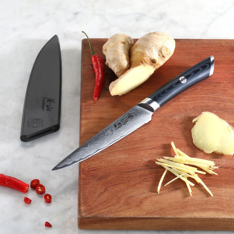 Serrated utility knife with patterned blade and black handle on wooden board with sliced ginger and red chili peppers, sheath