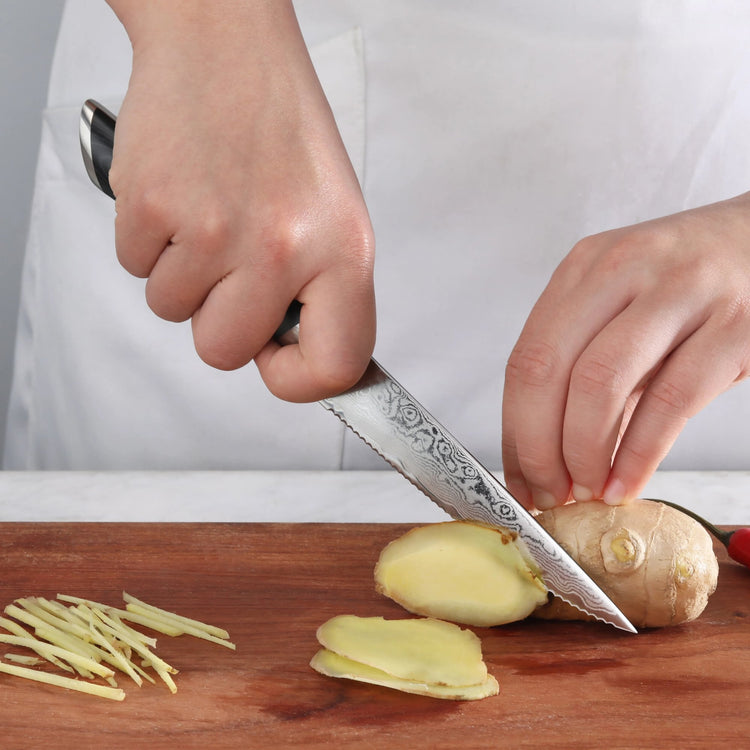 Hand holding serrated utility knife slicing ginger on wooden cutting board with thin ginger strips nearby.