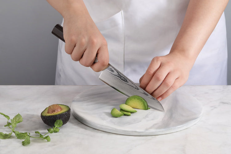 A person using the MAYA 5.5-inch Damascus steel prep utility knife to slice an avocado on a marble board.