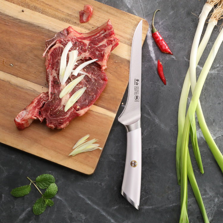 6-inch boning knife with white handle next to a wooden cutting board holding a raw steak topped with sliced onions.
