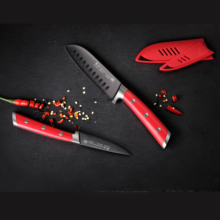 Two red-handled knives with black blades, a red chili pepper, chili pieces, and a red knife sheath on a black surface.