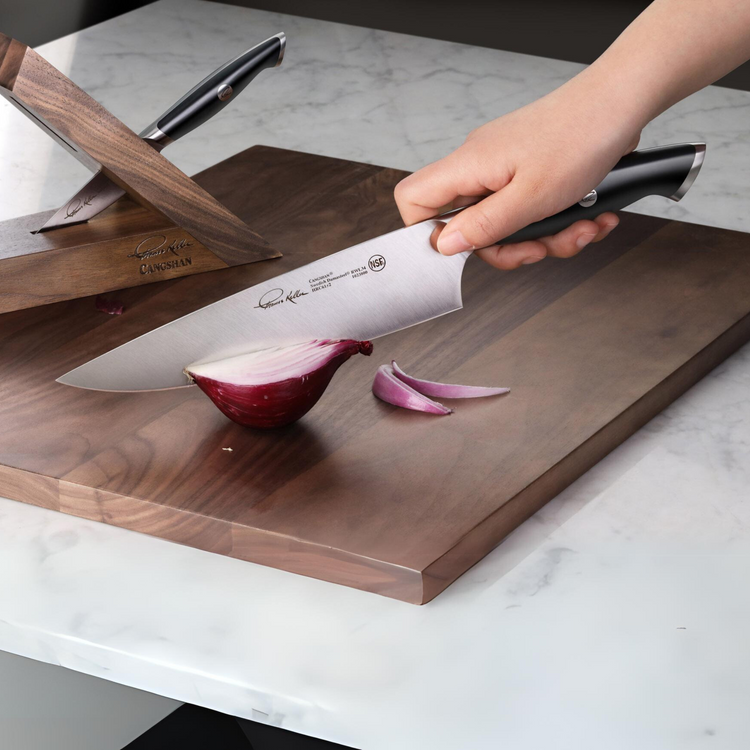 Hand holding a chef's knife slicing a red onion on a wooden cutting board with a wooden knife block in the background.