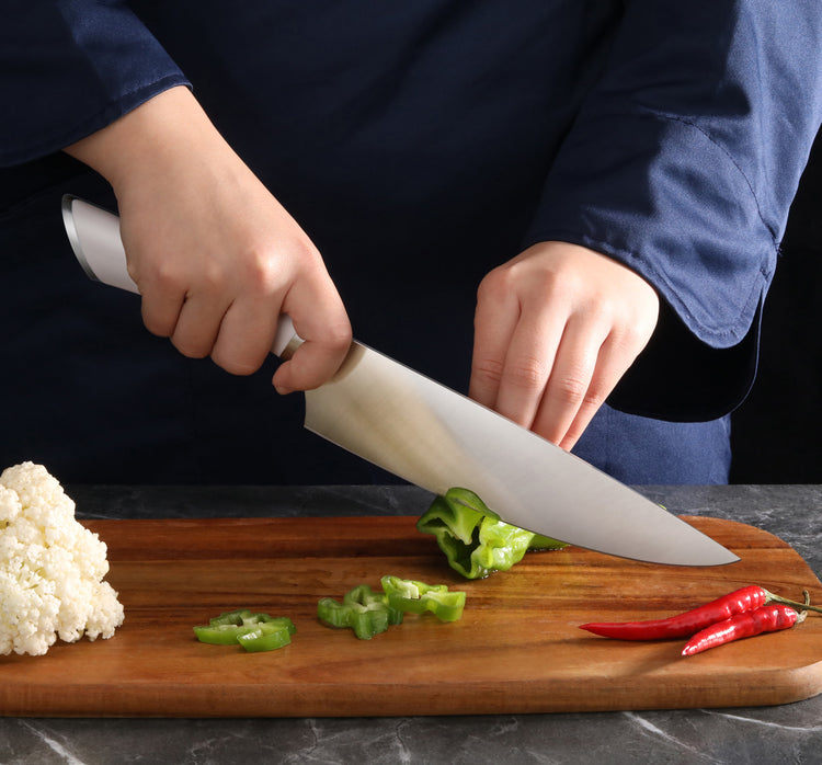 Hands slicing a green bell pepper on a wooden cutting board with cauliflower and red chili peppers nearby.