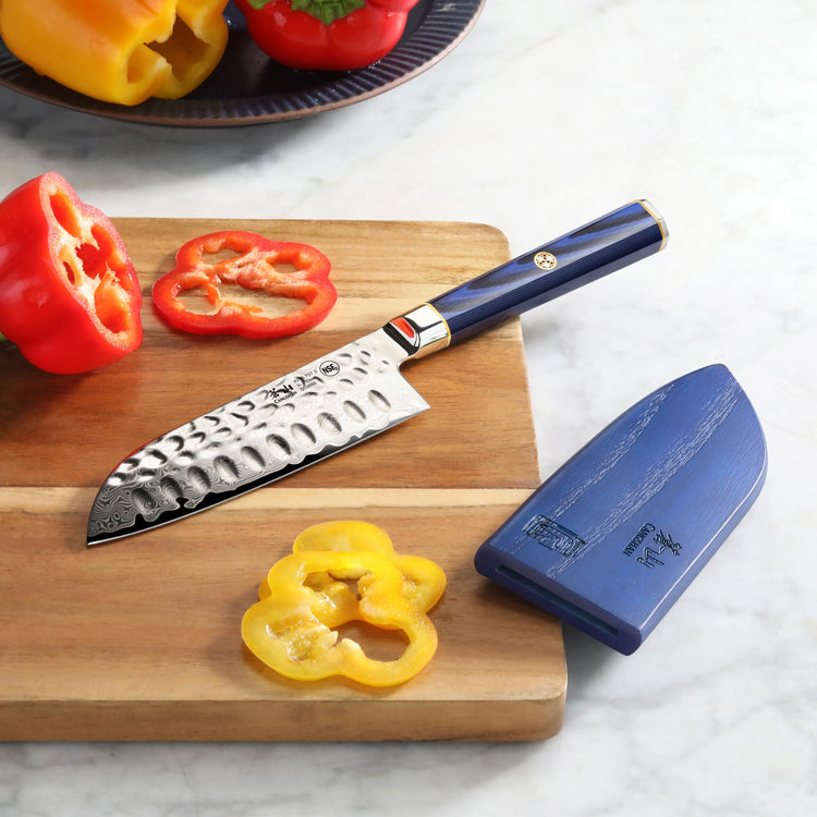 Santoku knife with hammered blade and blue handle on wooden cutting board with sliced red and yellow bell peppers and blue sh