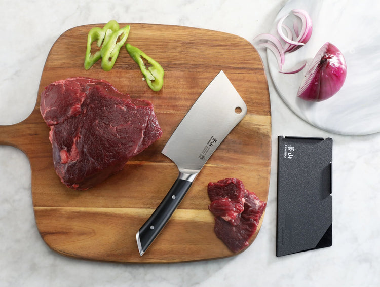 6-inch meat cleaver with black handle on wooden cutting board next to raw beef pieces and sliced green peppers.