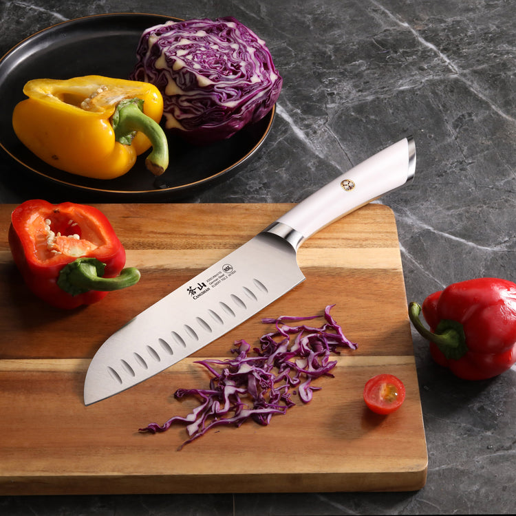 Santoku knife with white handle on wooden cutting board next to sliced purple cabbage, red bell pepper, and cherry tomato.