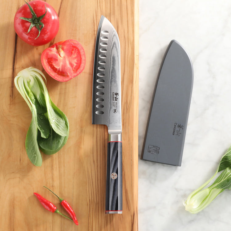 7-inch Santoku knife with patterned blade and black handle on wooden board with tomatoes, chili peppers, and sheath nearby.