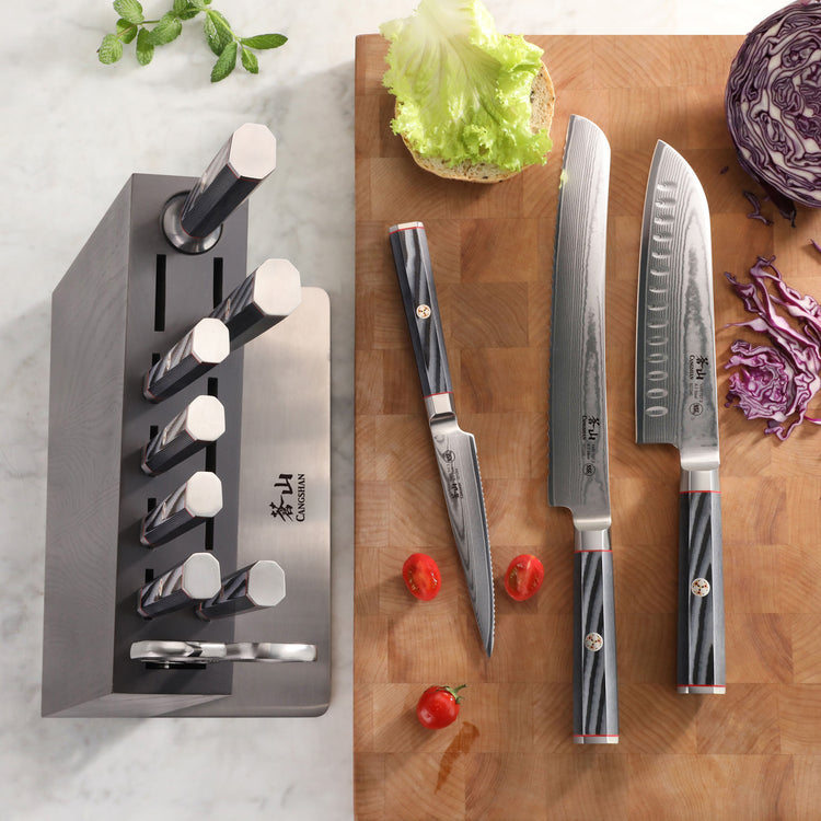 Knife block set with six knives in a black block and three knives laid out on a wooden cutting board with lettuce and cherry