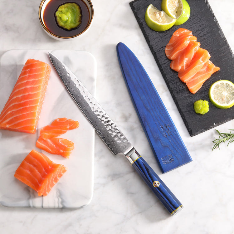 8-inch sashimi knife with blue sheath next to sliced salmon, lemon wedges, wasabi, and soy sauce on marble surface.