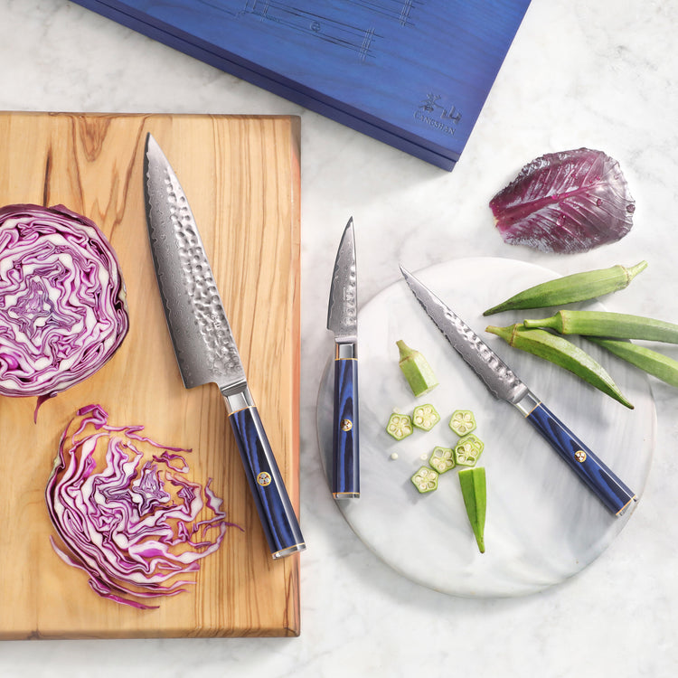 Three knives with blue handles on a wooden board and marble plate, next to sliced red cabbage and okra.
