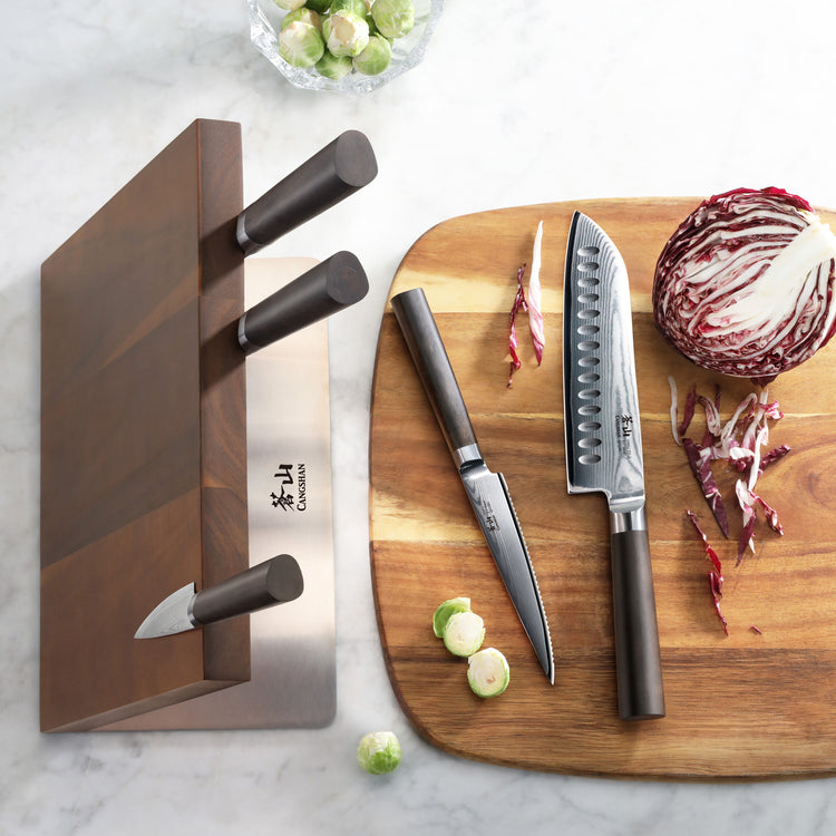 Six-piece knife set with dark handles in a wooden magnetic block and two knives on a wooden cutting board with radicchio.