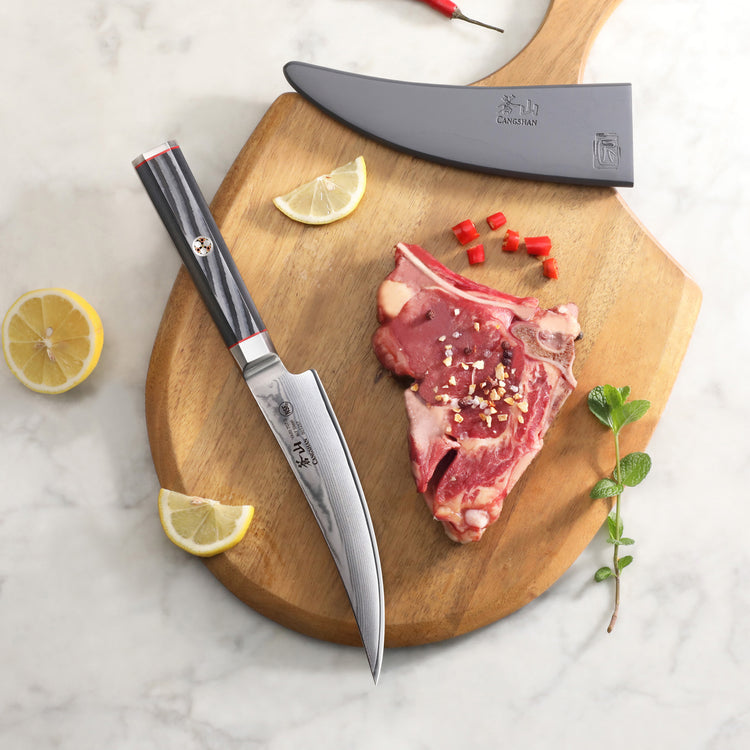6-inch boning knife with black handle and sheath on wooden board with raw meat, lemon wedges, and herbs.