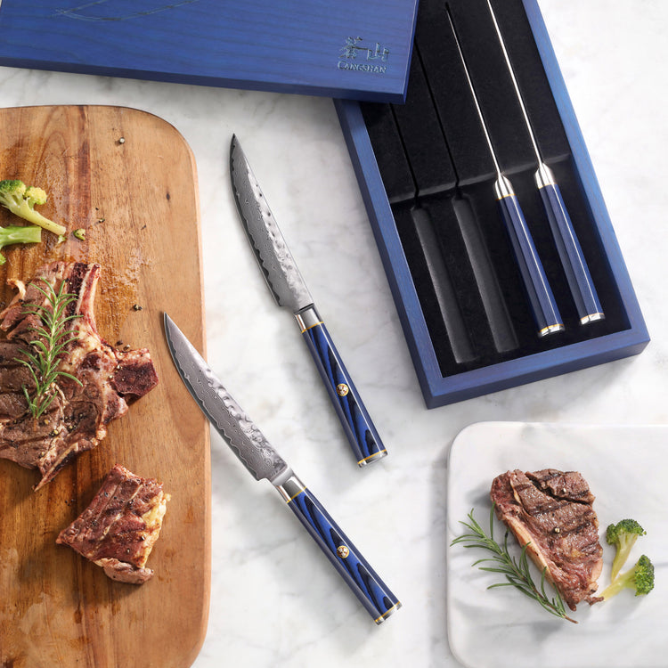 Four steak knives with blue handles and Damascus steel blades, two placed beside cooked steak on cutting boards.