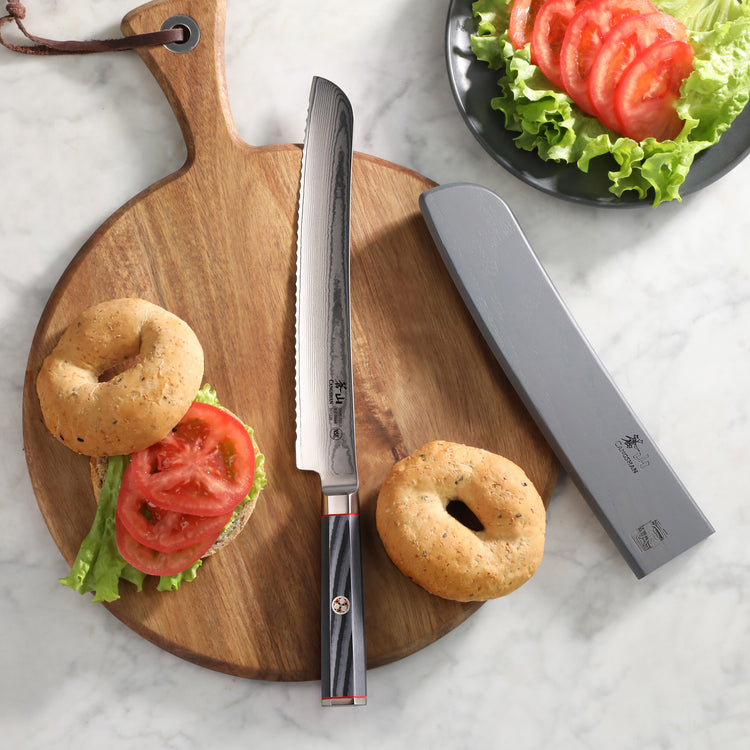 9-inch serrated bread knife with sheath on wooden cutting board, next to bagels and sliced tomatoes with lettuce.