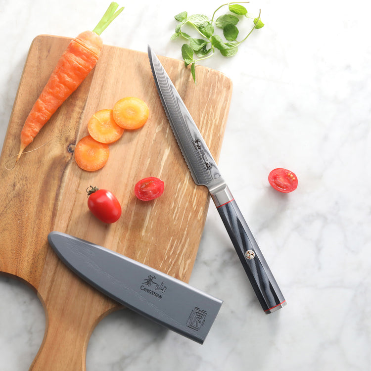 Serrated utility knife with sheath on wooden cutting board next to sliced carrot and cherry tomatoes.