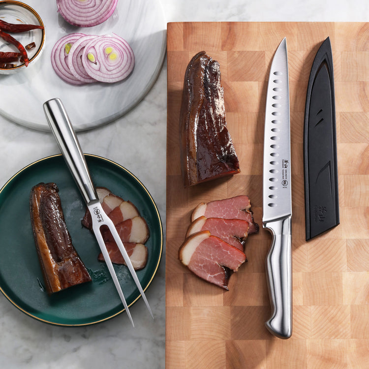 Two-piece carving set with a fork and a serrated knife with sheath, displayed with sliced meat and onion rings.