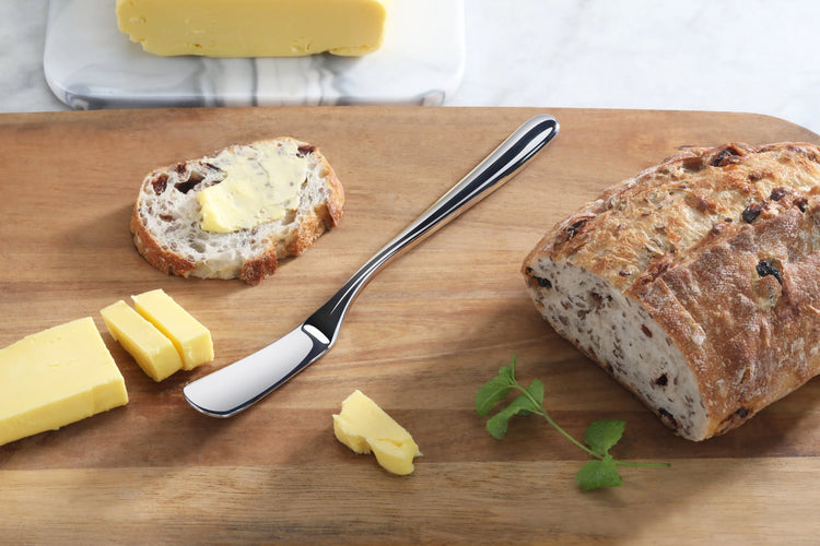 Butter knife with mirror finish on wooden board with sliced bread, butter, and herbs.