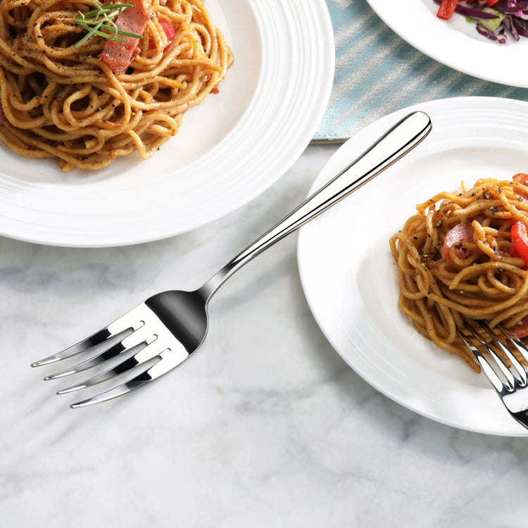 Serving fork with mirror finish placed on marble surface next to plates of spaghetti and salad.