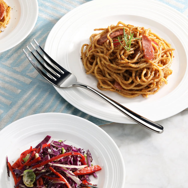 Serving fork with mirror finish placed beside a plate of spaghetti and a bowl of mixed vegetable salad.