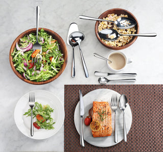 
                    
                      Slotted serving spoon with mirror finish resting in a wooden bowl of salad, alongside other flatware and plated food.
                    
                  