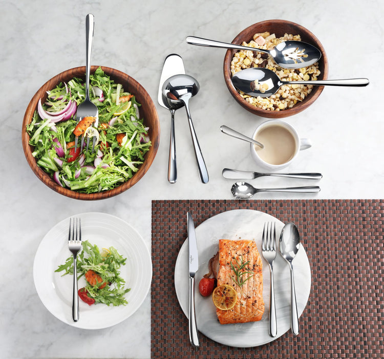 Slotted serving spoon with mirror finish resting in a wooden bowl of salad, alongside other flatware and plated food.