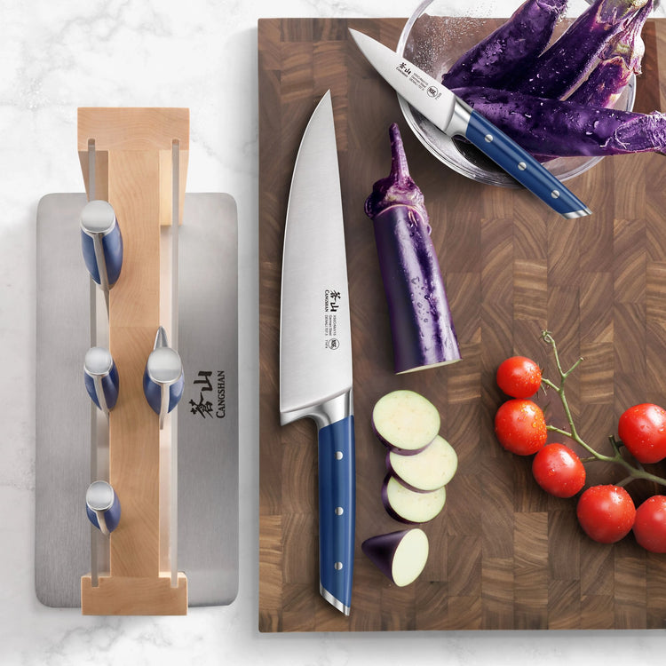 Seven-piece knife set with blue handles displayed on a wooden block and cutting board with sliced eggplant and cherry tomatoe