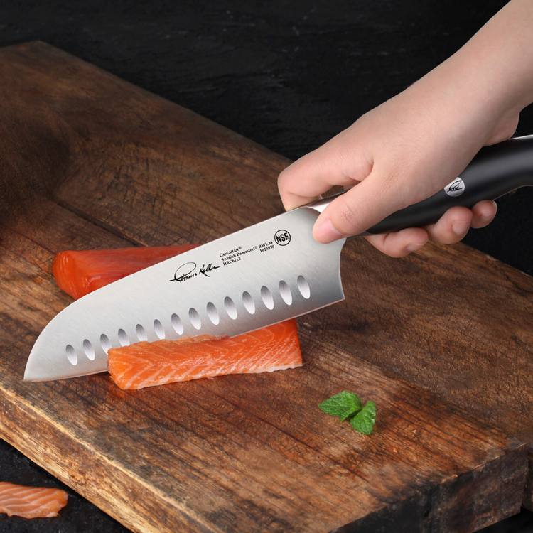 Hand holding TKSC 7-Inch Santoku Knife slicing raw salmon on a wooden cutting board with two mint leaves nearby.