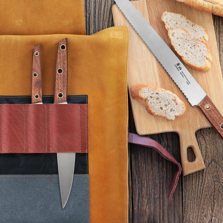 Two knives with wooden handles in a leather roll next to a serrated knife and sliced bread on a wooden cutting board.
