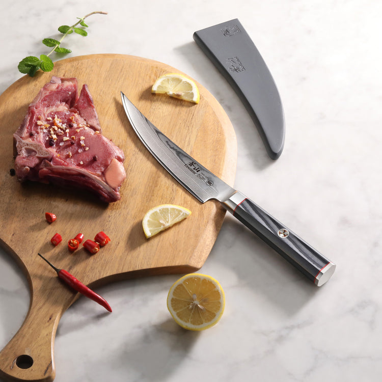 6-inch boning knife with polished handle and sheath next to raw meat, lemon slices, chili, and herbs on wooden board.