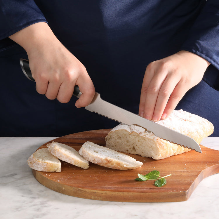 Hands slicing a loaf of bread on a wooden cutting board using a serrated offset bread knife.