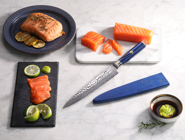 Blue-handled sashimi knife with sheath beside sliced raw and cooked salmon, lime, wasabi, and soy sauce on a marble surface.