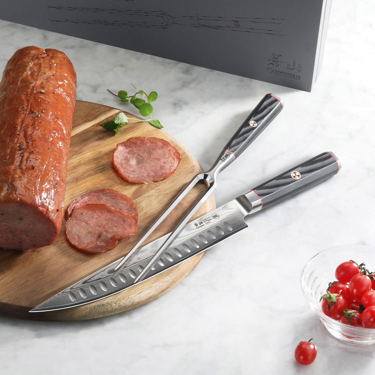 Two-piece carving set with black handles on a marble surface, next to sliced meat on a wooden board and a bowl of cherry toma
