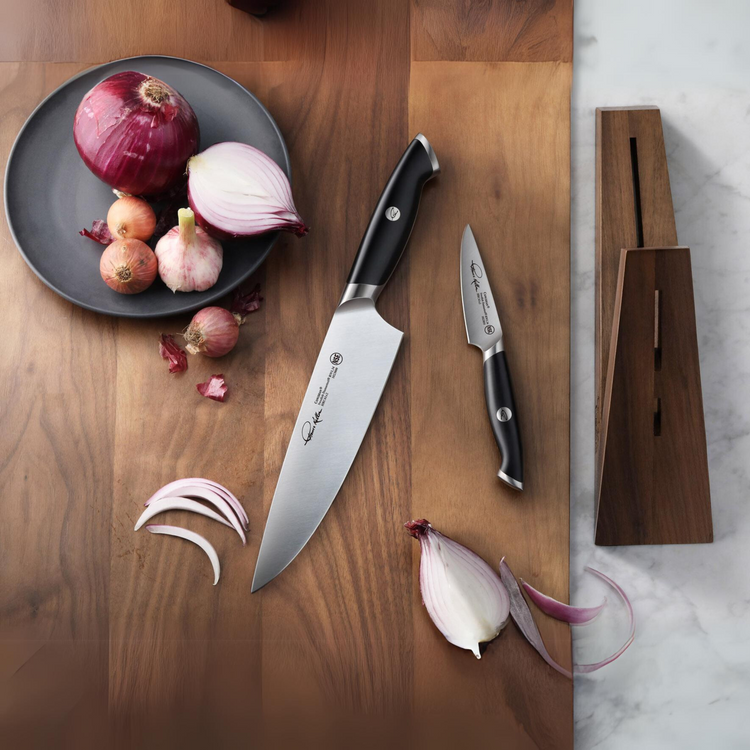 Two knives with black handles and engraved blades next to a wooden knife block, sliced onions, and whole onions on a wooden s