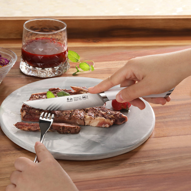 Hand holding ELBERT Series steak knife cutting a cooked steak on a round plate with a fork and glass of red beverage nearby.
