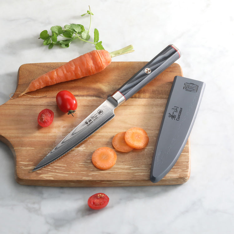Serrated utility knife with black handle and sheath on wooden board, alongside sliced carrot, whole carrot, and cherry tomato