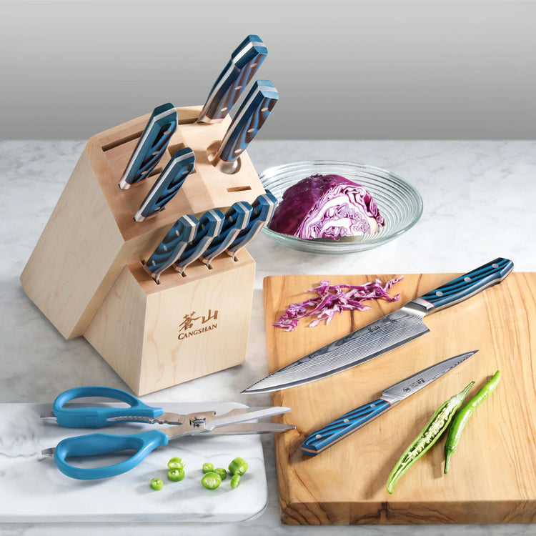 Wooden knife block with blue-handled knives, a pair of kitchen scissors, a cutting board with sliced green chili and two kniv