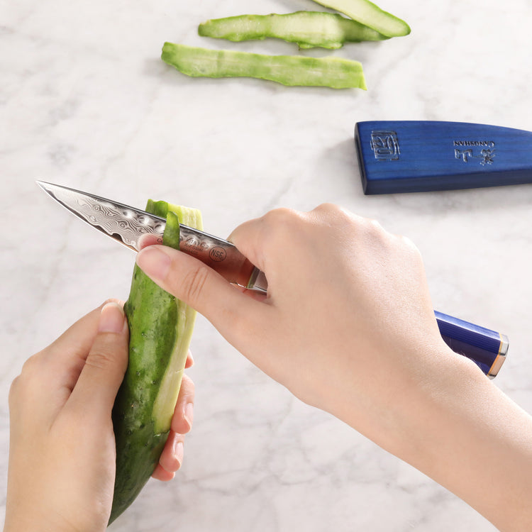 Hands peeling a cucumber with a small patterned knife and a blue sheath on a marble surface.