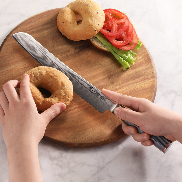 Hand holding a YARI Series 9-Inch Bread Knife with Sheath cutting a bagel on a wooden board with sandwich ingredients.