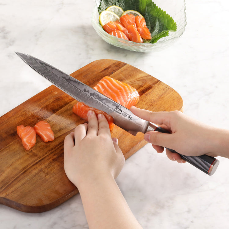 Hands slicing raw salmon on a wooden board with a 10-inch sashimi knife featuring a patterned blade and dark handle.