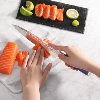 
                    
                      Hands slicing fresh salmon sashimi with a blue-handled knife on a white cutting board, with lemon and wasabi nearby.
                    
                  