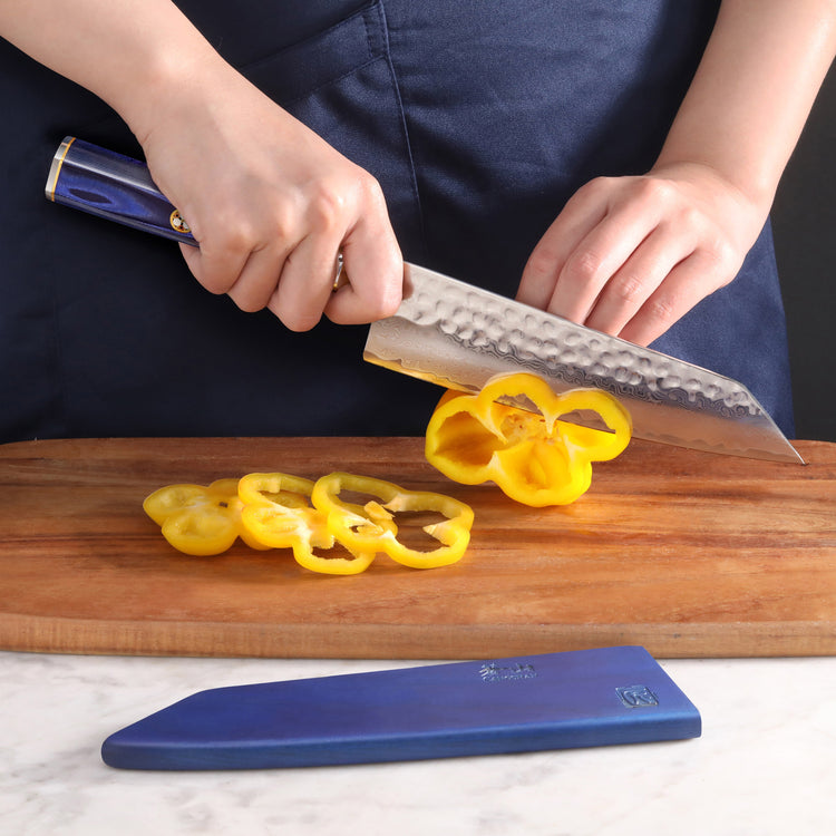 Hands slicing a yellow bell pepper on a wooden cutting board with a blue-handled Kiritsuke knife and blue sheath nearby.