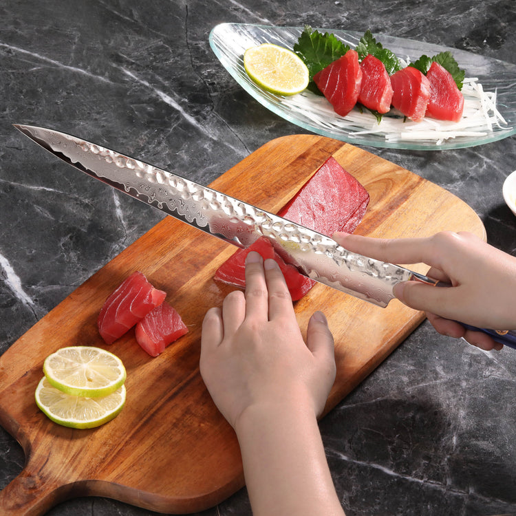 Hand holding a long, hammered steel sashimi knife slicing raw tuna on a wooden cutting board with lemon slices nearby.