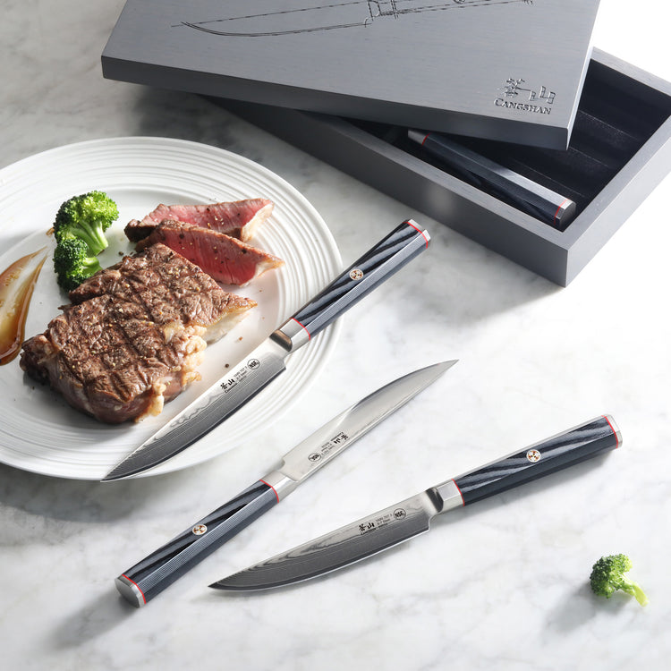 Three steak knives with black handles and Damascus steel blades displayed beside a plate with sliced steak and broccoli, with