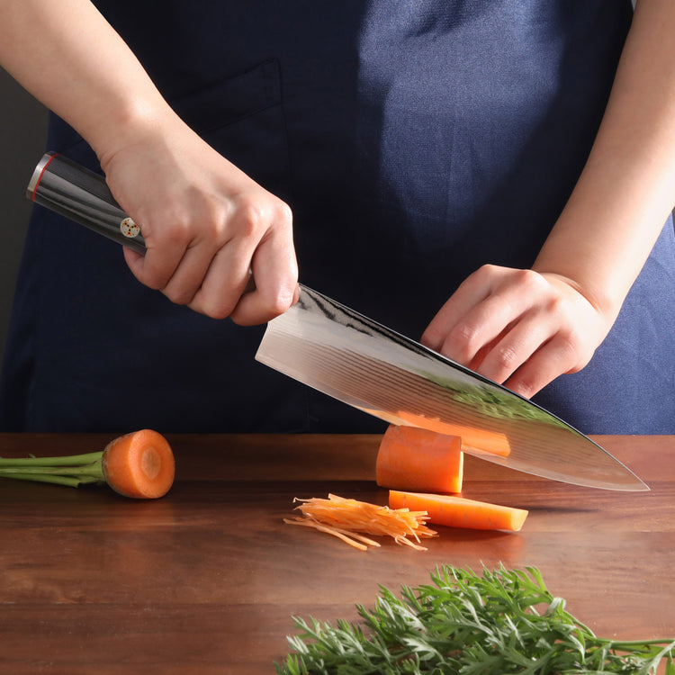 Hands using a large kitchen knife to slice a carrot on a wooden cutting board with carrot greens nearby.