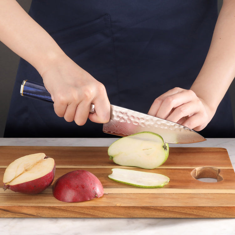 Hands using a blue-handled knife to slice a green apple on a wooden cutting board with apple pieces nearby.