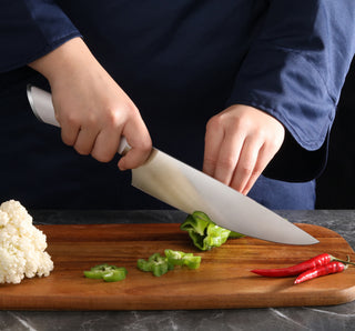 
                    
                      Hands slicing a green bell pepper on a wooden cutting board with a large chef's knife and red chili peppers nearby.
                    
                  