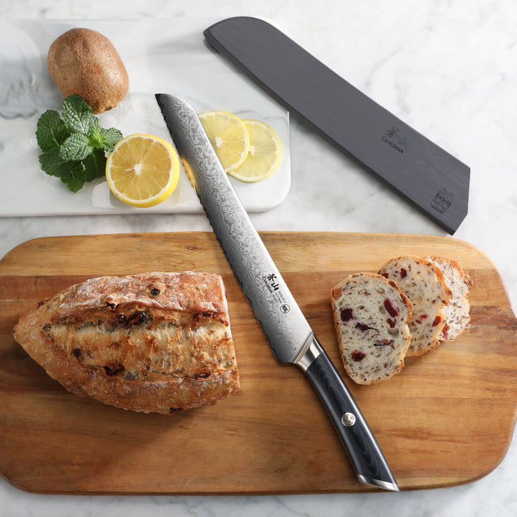 Bread knife with Damascus steel blade and black handle on wooden board with sliced bread, lemon, mushroom, and herbs.