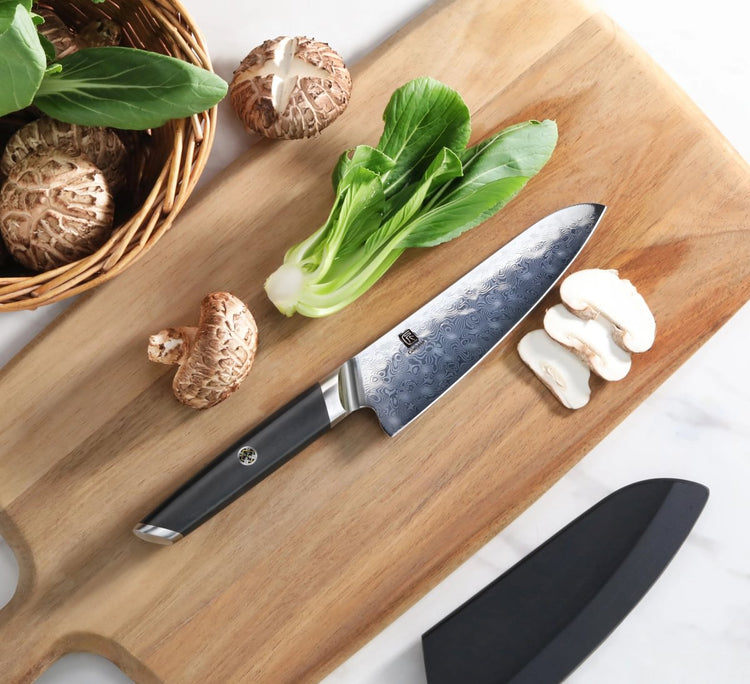 Patterned steel utility knife on a wooden cutting board with bok choy, sliced mushrooms, and a black sheath.