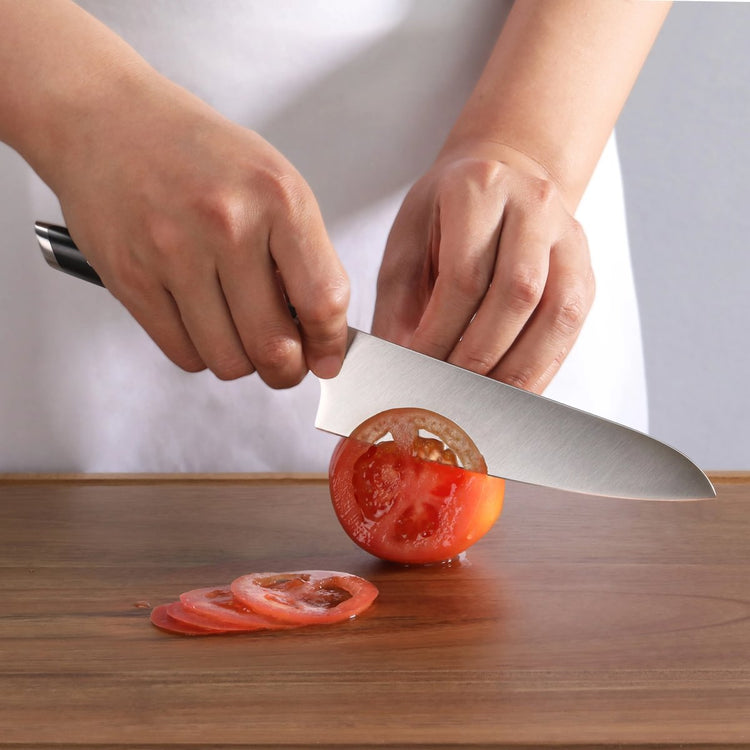 Hands slicing a tomato with a large kitchen knife on a wooden cutting board.
