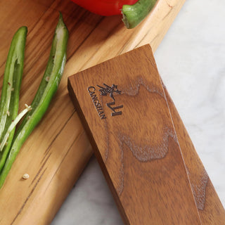 
                    
                      Ash wood magnetic knife sheath with engraved logo, placed next to green chili peppers on a wooden cutting board.
                    
                  